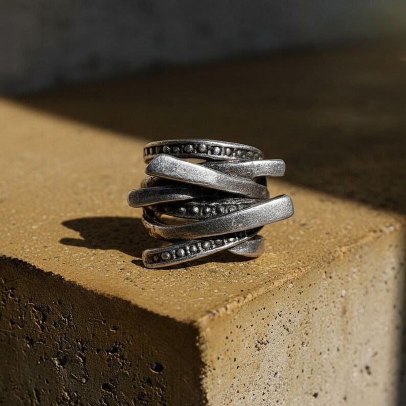 Oxidized silver ring with layered overlapping bands and dotted metal detailing, placed on a rough stone surface in natural sunlight. The design shows a mix of matte and darkened finishes with sculptural depth. Contemporary handcrafted jewelry by Rannka with industrial edge.