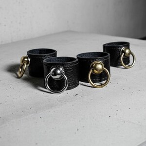 Set of black vegan leather rings with metal sphere and loop hardware in silver and antique brass finishes, arranged on a concrete surface. Minimal industrial composition showing texture contrast and material quality of the modern unisex jewelry design.
