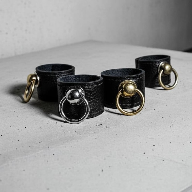 Set of black vegan leather rings with metal sphere and loop hardware in silver and antique brass finishes, arranged on a concrete surface. Minimal industrial composition showing texture contrast and material quality of the modern unisex jewelry design.