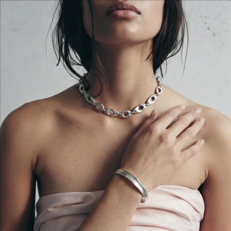 Close-up of a model wearing a chunky silver chain necklace with oval links and a matching minimal silver cuff bracelet. The jewelry is styled against bare skin with a soft blush fabric wrap, neutral background, and natural light, creating a clean, modern, sculptural look aligned with Rannka’s architectural aesthetic.