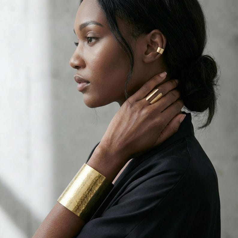 Modern sculptural gold ring worn on the hand, paired with a minimalist gold ear cuff and wide cuff bracelet, styled on a model in a clean editorial setting with soft natural light.