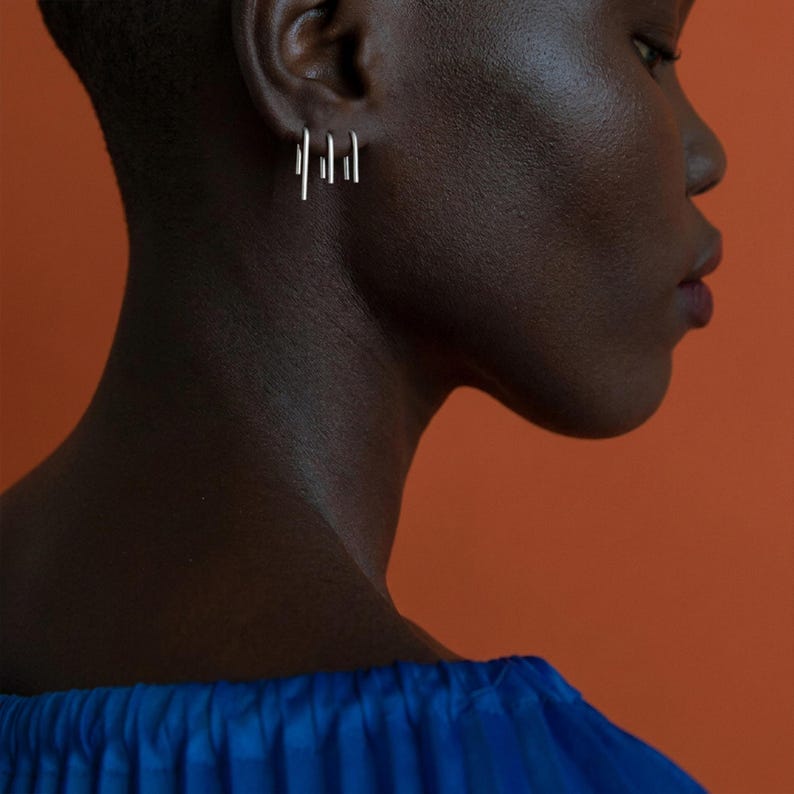 Sterling silver slip earrings in small and medium lengths styled as an asymmetric ear stack, minimalist bar drop earrings on ear, modern sculptural jewelry look, clean industrial aesthetic, subtle everyday silver earrings, close-up side profile with warm orange backdrop, Rannka style, refined and editorial mood