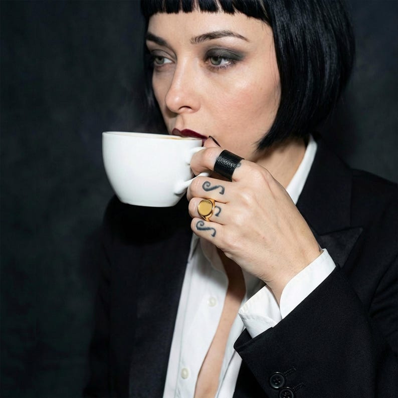 woman with short black bob haircut and dark lipstick holding a white cup close to her face, wearing a black blazer over a white shirt, visible hand tattoos and black nail polish, wearing a 14k gold round signet ring and a wide black band ring, close-up portrait with soft lighting and dark background