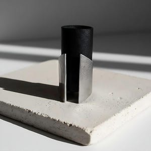 Black vegan leather and stainless steel statement ring displayed on a textured white stone block. The design features a tall leather band with brushed steel edges forming an open architectural shape. Minimalist product photography with sharp shadows and modern industrial lighting.