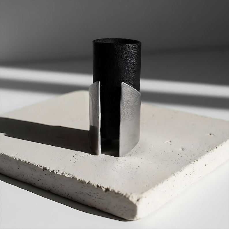 Black vegan leather and stainless steel statement ring displayed on a textured white stone block. The design features a tall leather band with brushed steel edges forming an open architectural shape. Minimalist product photography with sharp shadows and modern industrial lighting.