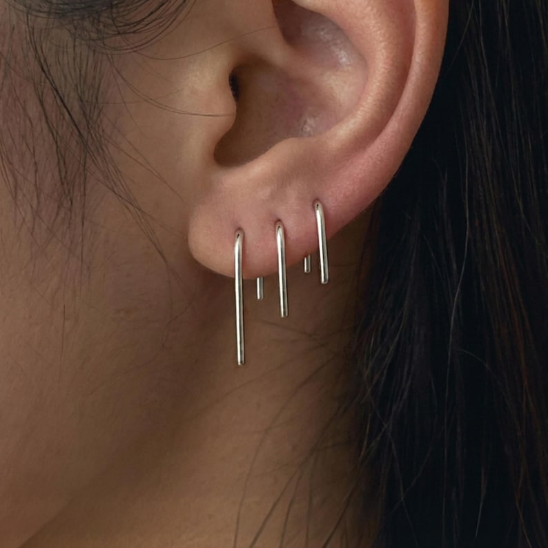 Mini slip earrings in sterling silver shown on ear, minimalist thin bar drop earrings with clean vertical lines, modern everyday jewelry, delicate silver ear stack styling, subtle industrial aesthetic, Rannka style, close-up ear detail with multiple piercings, sleek and understated mood