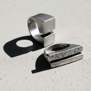 Two silver statement rings with geometric forms — one featuring a split cube design, the other a hammered layered structure. Photographed in sharp sunlight to highlight their sculptural texture. Brutalist minimalist jewelry by Rannka.
