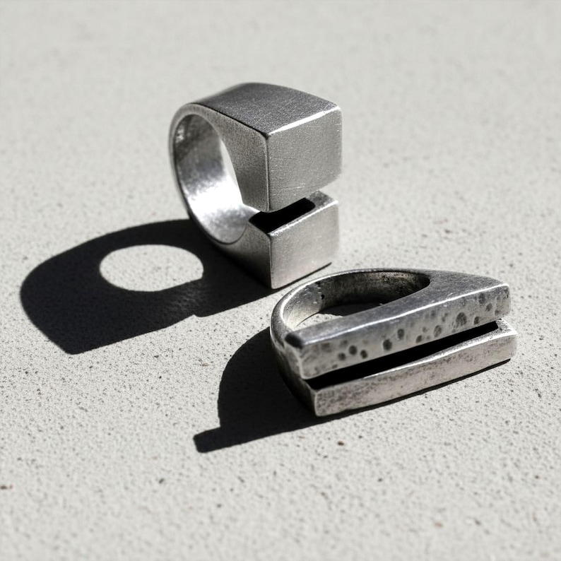 Two silver statement rings with geometric forms — one featuring a split cube design, the other a hammered layered structure. Photographed in sharp sunlight to highlight their sculptural texture. Brutalist minimalist jewelry by Rannka.