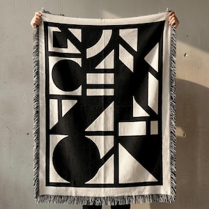 Black and cream woven brutalist blanket with bold geometric shapes, including circles, triangles, lines, and blocks arranged in a graphic abstract layout. High-contrast pattern framed by a light border, finished with fringe edges. Blanket is held up against a raw concrete wall, highlighting its architectural, modern, and minimalist design. Textured woven fabric with a strong visual structure and clean, graphic presence. Rannka style.