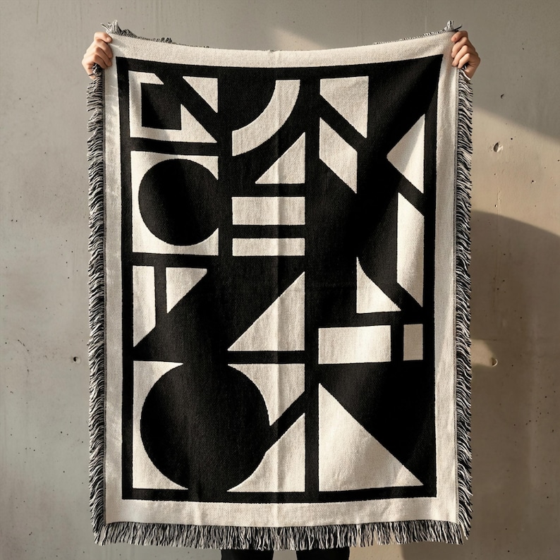 Black and cream woven brutalist blanket with bold geometric shapes, including circles, triangles, lines, and blocks arranged in a graphic abstract layout. High-contrast pattern framed by a light border, finished with fringe edges. Blanket is held up against a raw concrete wall, highlighting its architectural, modern, and minimalist design. Textured woven fabric with a strong visual structure and clean, graphic presence. Rannka style.