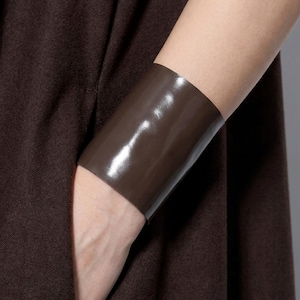 May include: A wide, smooth, dark brown cuff bracelet with a glossy, reflective finish. The bracelet is solid in color and features a simple, minimalist band design that wraps around the wrist, contrasting with the dark brown fabric garment.