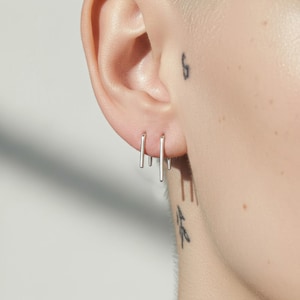 Sterling silver slip hoop earrings in graduated lengths, modern arc wire earrings styled across multiple piercings, sleek curved bar earrings with tapered ends, minimalist ear layering, lightweight silver hoops for everyday wear, close-up ear with fine line tattoos, clean sculptural jewelry, Rannka style