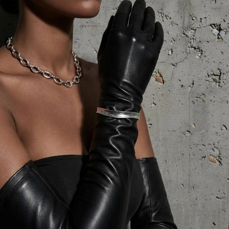 Editorial close-up of a model wearing a sculptural silver oval-link chain necklace and a minimal silver cuff bracelet, styled with long black leather gloves and a strapless black top. Shot against a raw concrete wall with dramatic directional light, highlighting metal texture, contrast, and a bold architectural mood consistent with Rannka’s aesthetic.
