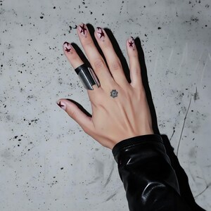 Hand wearing a silver and black ring made from metal and vegan leather, photographed on a textured concrete surface. The ring’s angular form contrasts with the sleek leather sleeve and star nail art, giving it a bold, modern edge.