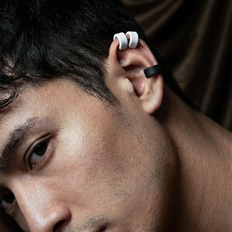 Black and white ear cuff stack worn on upper ear and mid ear, no piercing ear jewelry styled on a male model, matte ear wraps hugging the ear rim, modern unisex ear accessories with clean graphic contrast, close-up portrait detail by Rannka.