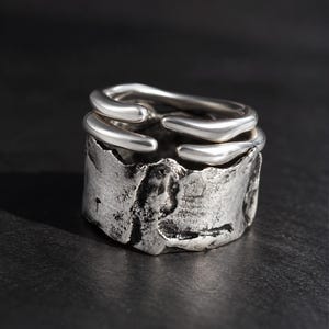 May include: A silver ring featuring a wide, textured band with a dark, weathered finish and a smooth, polished upper section consisting of two curved, open-ended bands that wrap around the top of the ring in a layered, organic design.