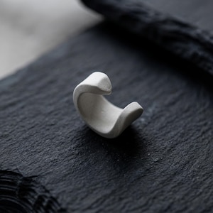 White ear cuff shown as a single piece on dark textured stone, no piercing ear jewelry with open curved form, sculptural minimalist ear wrap for modern styling, matte finish ear accessory designed for stacking or solo wear, contemporary Rannka ear jewelry.