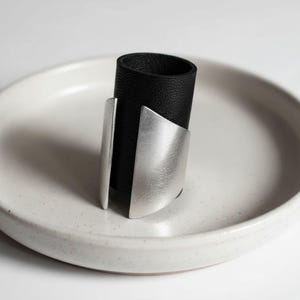 Black vegan leather and matte silver ring displayed on a white ceramic dish. The ring features a tall band with curved metallic plates on each side, creating a clean geometric silhouette. Soft natural light and neutral tones emphasize the modern, minimalist jewelry design.