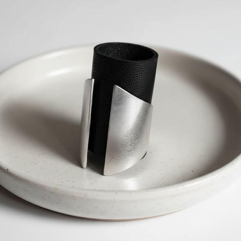 Black vegan leather and matte silver ring displayed on a white ceramic dish. The ring features a tall band with curved metallic plates on each side, creating a clean geometric silhouette. Soft natural light and neutral tones emphasize the modern, minimalist jewelry design.