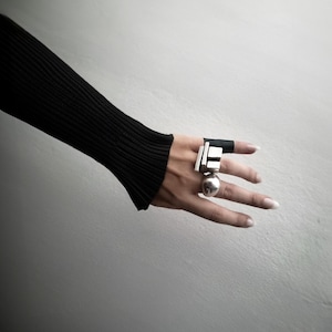 Hand styled with bold sculptural rings — a geometric bar ring, a domed silver sphere ring, and a black band ring. Worn with a ribbed black sleeve for a modern, architectural look. Avant-garde jewelry styling by Rannka.
