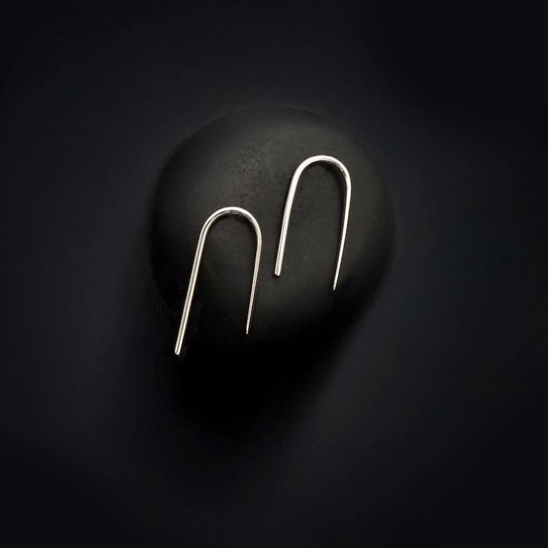 Minimalist silver arc slip earrings by Rannka displayed on a matte black round surface. The curved design is clean and modern, photographed in moody lighting that highlights their dainty form. Ideal for everyday wear or as a gift for her, this earring set works well for second hole styling or worn alone for a subtle, contemporary look.