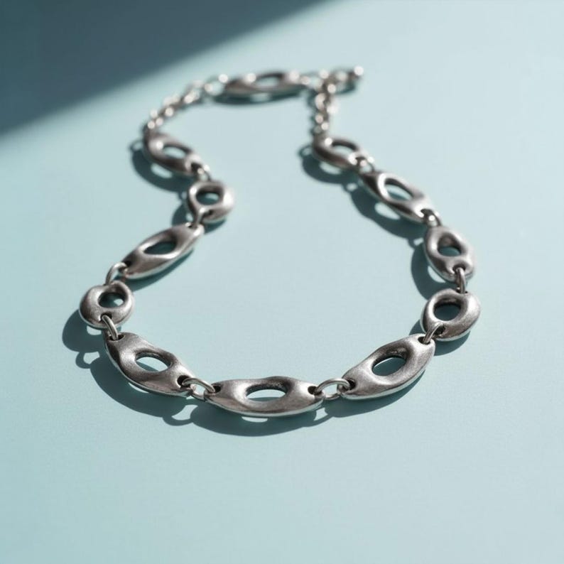 Close-up of a sculptural silver necklace with irregular open oval links displayed on a soft blue surface. The design has an organic, modern-art feel with smooth contours and a slightly industrial edge, catching light to reveal its fluid metallic texture.