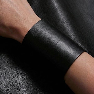Può includere: Un bracciale a polsino in pelle nera. L'ampia fascia è realizzata in un materiale di pelle strutturata. Il polsino è indossato al polso e ha un design semplice e minimalista. Il bracciale è di colore nero.