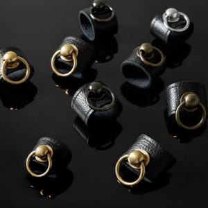 Collection of black vegan leather rings with metal sphere and loop accents in gold, brass, gunmetal, and silver finishes. Arranged on reflective black surface, showcasing texture, shine, and modern minimalist craftsmanship of the unisex jewelry pieces.
