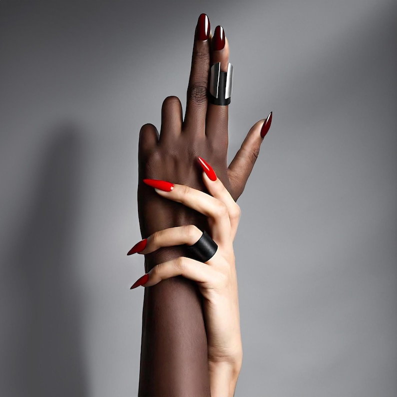 Two hands intertwined, showcasing modern sculptural rings made of matte black vegan leather and brushed steel. The design has a sharp architectural edge, photographed with studio lighting against a neutral gray backdrop. Red polished nails add contrast and a high-fashion editorial feel to the minimal composition.