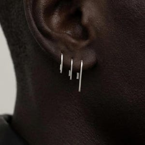 May include: Three silver earrings are displayed in a person's ear. Each earring has a minimalist design, with a straight bar and a few short lines. The earrings are small and delicate, adding a subtle touch of style.