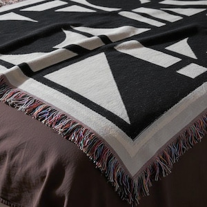 Close-up of a black and cream geometric woven blanket with a bold angular pattern and multicolor fringed edges. The texture is tight and structured, draped neatly over a brown bed. Modern brutalist-style home textile by Rannka, blending art and functional decor.