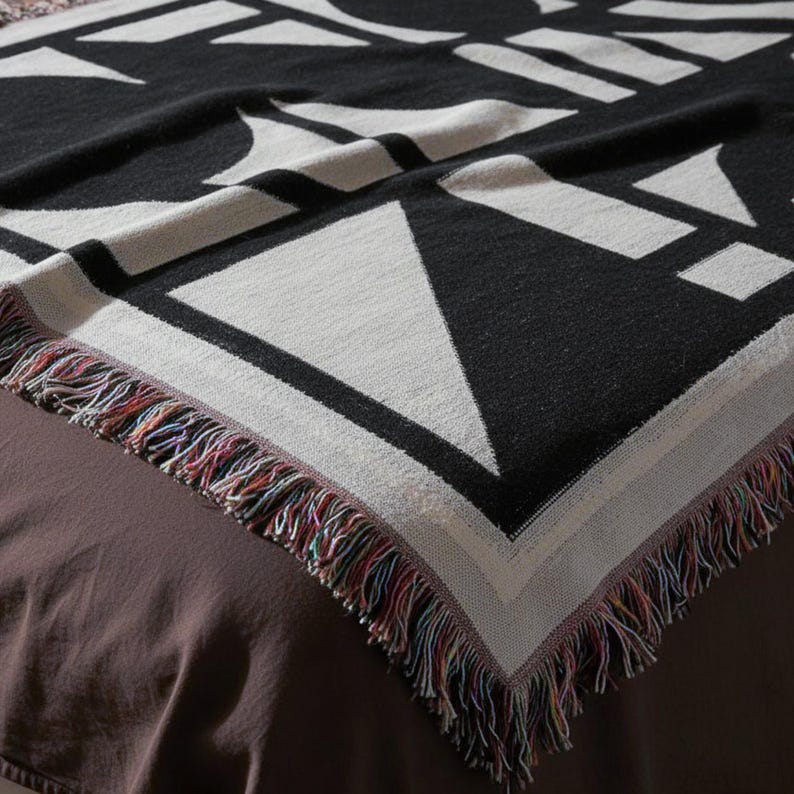 Close-up of a black and cream geometric woven blanket with a bold angular pattern and multicolor fringed edges. The texture is tight and structured, draped neatly over a brown bed. Modern brutalist-style home textile by Rannka, blending art and functional decor.