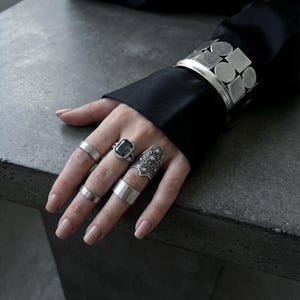 May include: A hand displaying a collection of silver rings and a cuff bracelet. The rings include a large square-cut gemstone ring, a detailed ornate ring, and several simple bands. The cuff bracelet features a geometric design.
