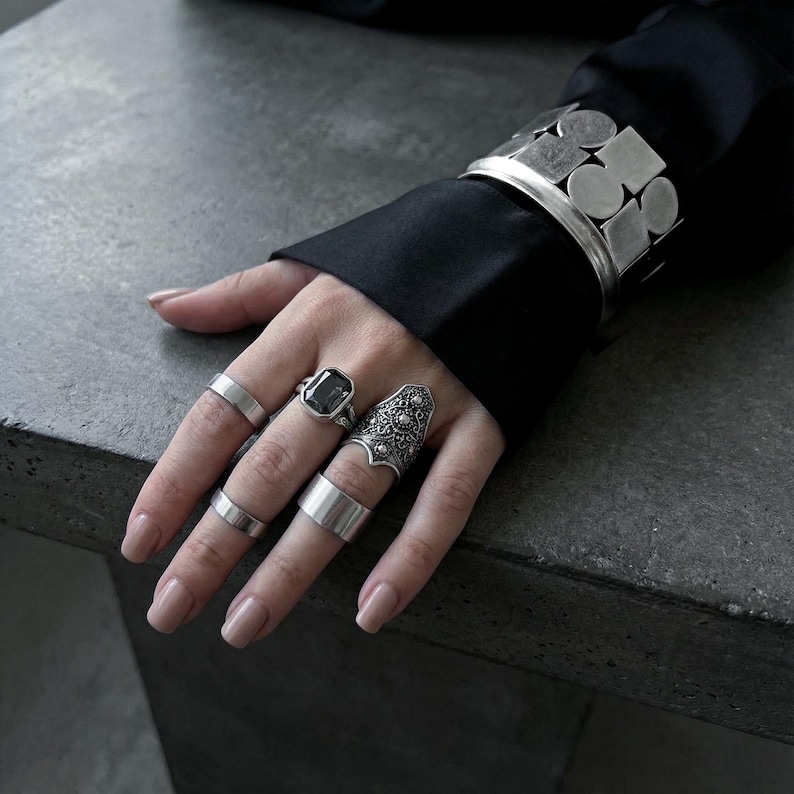 May include: A hand displaying a collection of silver rings and a cuff bracelet. The rings include a large square-cut gemstone ring, a detailed ornate ring, and several simple bands. The cuff bracelet features a geometric design.