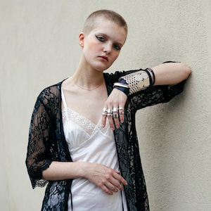 Model wearing silver statement rings and a wide metal and leather cuff bracelet, styled with a white slip dress and black lace robe. Close-up fashion portrait with short hair, minimal makeup, natural light, and a plain textured wall background. Rannka styling.