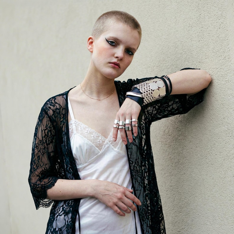 Model wearing silver statement rings and a wide metal and leather cuff bracelet, styled with a white slip dress and black lace robe. Close-up fashion portrait with short hair, minimal makeup, natural light, and a plain textured wall background. Rannka styling.