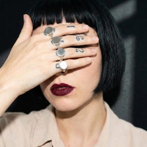 woman with short black bob covering her eyes with her hand, wearing multiple stacked 925 silver round signet rings across fingers, dark red lipstick, neutral beige shirt, close-up portrait on dark background