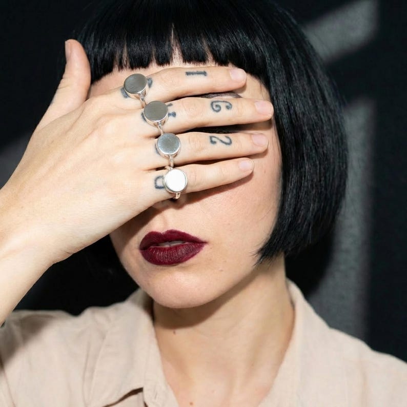 woman with short black bob covering her eyes with her hand, wearing multiple stacked 925 silver round signet rings across fingers, dark red lipstick, neutral beige shirt, close-up portrait on dark background
