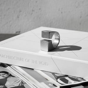 Open silver cube ring placed on a book titled *Architecture of the Void*. The minimalist matte surface and geometric form create a sculptural, design-focused mood. Modern art-inspired unisex jewelry by Rannka.
