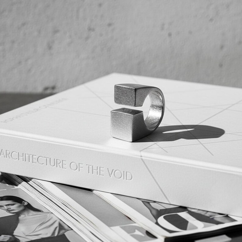 Open silver cube ring placed on a book titled *Architecture of the Void*. The minimalist matte surface and geometric form create a sculptural, design-focused mood. Modern art-inspired unisex jewelry by Rannka.