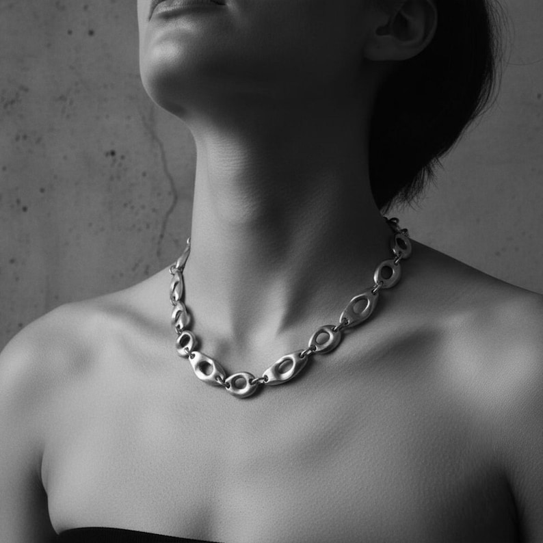 Black and white close-up of a woman wearing a sculptural silver chain necklace with smooth oval links. The minimalist composition highlights the jewelry’s bold yet refined form, creating a timeless, architectural look against bare skin.