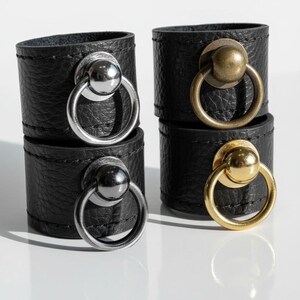 Close-up of four black vegan leather rings featuring round metal sphere and loop hardware in silver, gold, and antique brass finishes. Displayed under natural light on a reflective white surface, highlighting the contrast between matte leather texture and polished metal detail.