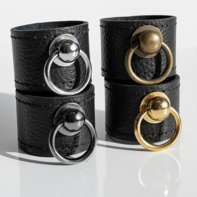 Close-up of four black vegan leather rings featuring round metal sphere and loop hardware in silver, gold, and antique brass finishes. Displayed under natural light on a reflective white surface, highlighting the contrast between matte leather texture and polished metal detail.