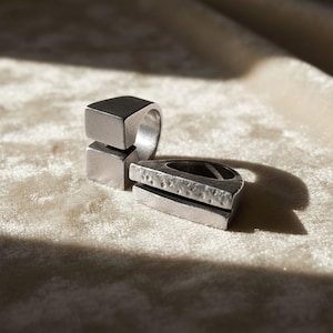 Two contemporary silver rings resting on soft beige fabric under natural light. One features a square open design, the other a textured geometric bar shape. Artisan-made modern jewelry for everyday wear, art-inspired accessories by Rannka.
