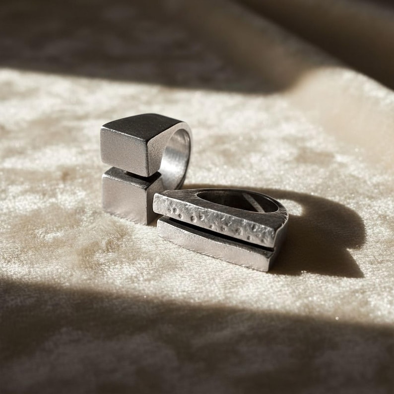 Two contemporary silver rings resting on soft beige fabric under natural light. One features a square open design, the other a textured geometric bar shape. Artisan-made modern jewelry for everyday wear, art-inspired accessories by Rannka.