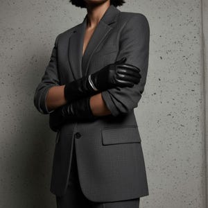 Model in a tailored gray suit with sleeves rolled up, wearing black leather gloves and a silver cuff bracelet. The composition emphasizes structure, contrast, and quiet strength — polished tailoring meets raw metal detail. A minimalist portrait of modern power dressing.
