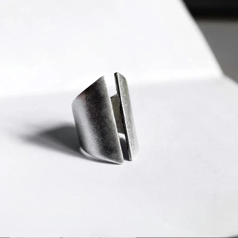 Modern adjustable metal ring with a sculptural split-front design, matte brushed finish, and raw industrial look. Open band construction allows a flexible fit. Minimal, architectural style jewelry photographed on a clean white background with soft shadows. Contemporary unisex ring by Rannka, focused on form, texture, and bold negative space.