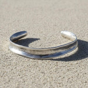 Silver cuff bracelet photographed on sand under natural light. The sunlight reveals fine surface details, subtle dents, and organic shaping. The composition feels raw and elemental, blending jewelry craftsmanship with an earthy, minimal setting.