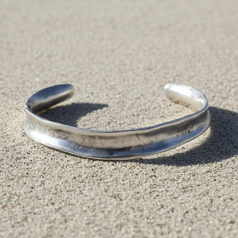 Silver cuff bracelet photographed on sand under natural light. The sunlight reveals fine surface details, subtle dents, and organic shaping. The composition feels raw and elemental, blending jewelry craftsmanship with an earthy, minimal setting.