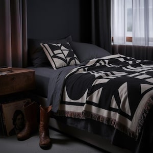 Black and cream geometric woven blanket with multicolor fringed edges styled on a dark-toned bed with a matching pillow. The dimly lit room features wooden accents, leather boots, and a candle, creating a moody, modern interior atmosphere.